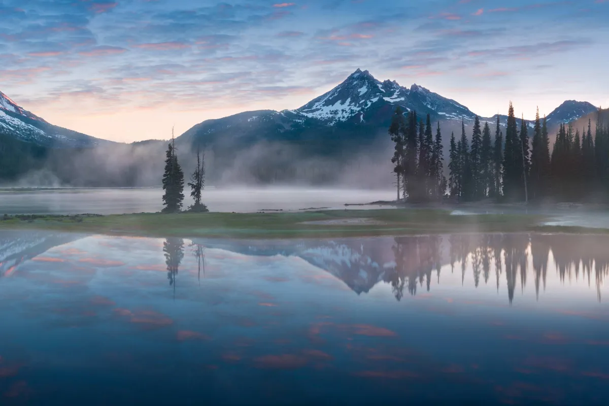 A tranquil view of a serene lake bordered by trees with mountains in the distance. The sky reflects on the calm water, creating a soothing and picturesque scene ideal for relaxation, hiking, or quiet reflection.