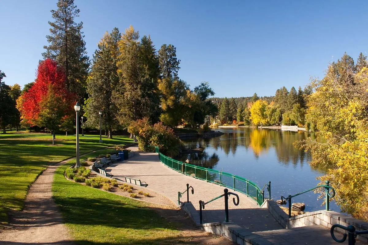 A tranquil riverside path lined with colorful trees and a green railing, the calm water reflecting the sky’s soft hues. The path invites leisurely strolls, and the surrounding natural beauty offers a peaceful escape just steps from the heart of Redmond.