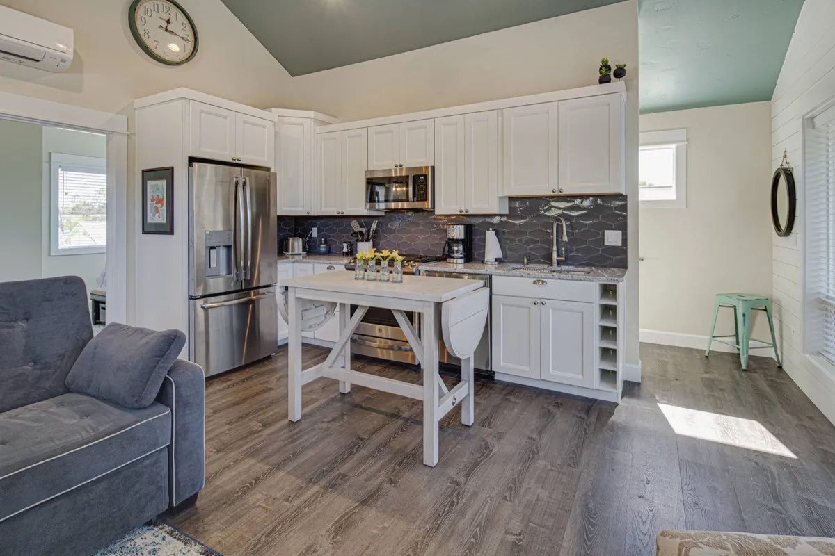 A kitchen island takes center stage, surrounded by stainless steel appliances and illuminated by natural light streaming through a nearby window. A decorative clock and thoughtful details create a cozy, welcoming mood perfect for both casual cooking and socializing.
