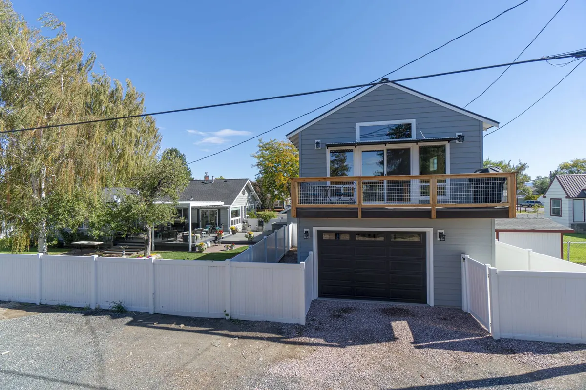 A modern house facade featuring a welcoming balcony and a fenced yard, surrounded by greenery. The house looks inviting, with space for outdoor gatherings, morning coffee on the porch, and evening strolls.