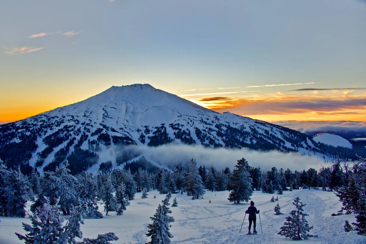 A snow-covered mountain looms under a vibrant sunset sky, with a silhouetted skier in the midground and pine trees dotting the landscape. The scene is both adventurous and serene, evoking the thrill of winter sports and the beauty of Central Oregon’s natural surroundings.