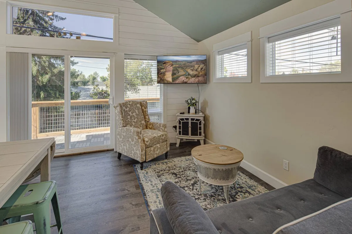 This image presents a comfortable living area featuring a plush, patterned armchair, a flat-screen television showing scenic imagery, a round coffee table, and large windows that allow an abundance of natural light to fill the room. The overall ambiance is serene and inviting, aligning with the description of a sun-drenched, relaxing living space.