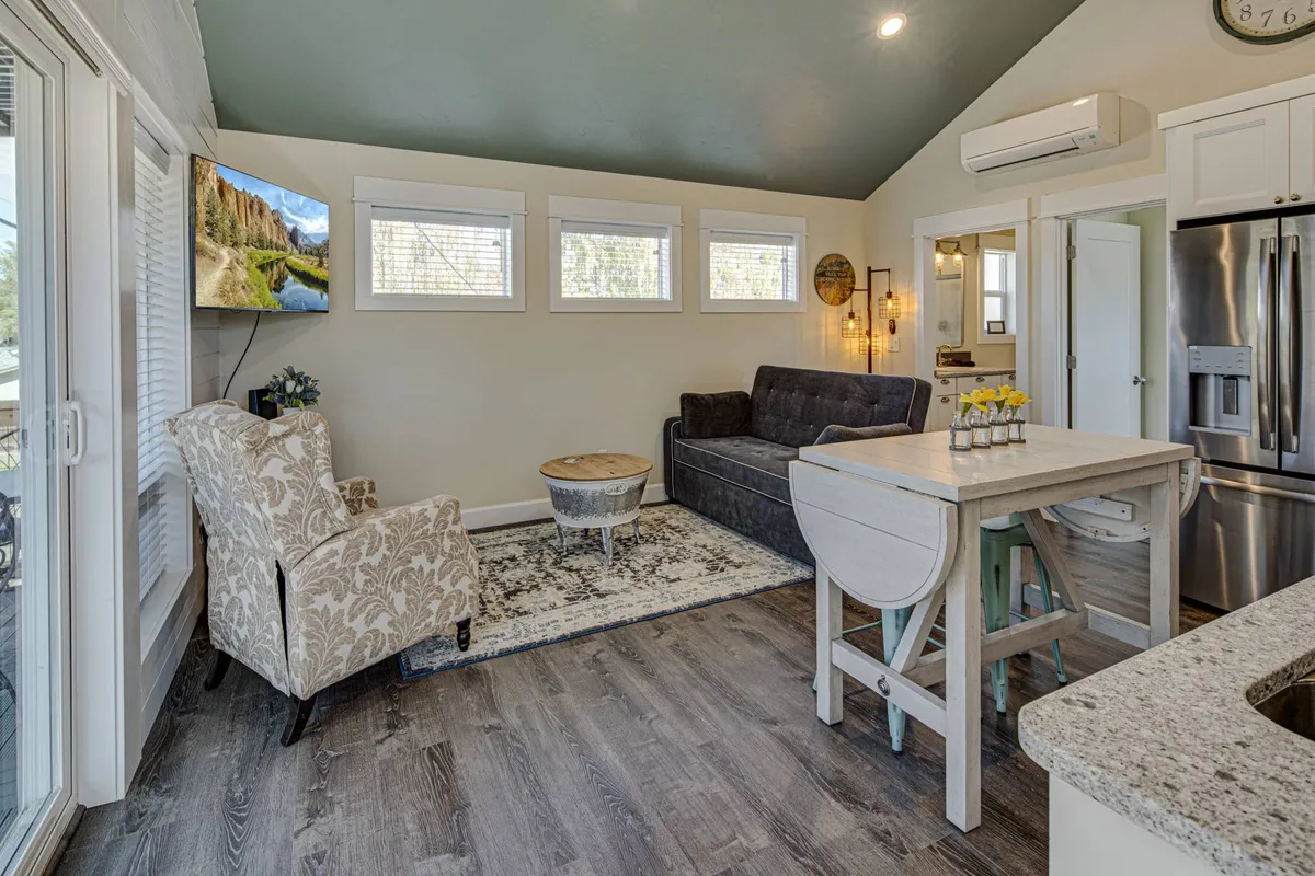 The photo displays a homey living area with a comfy seating arrangement, a kitchen counter visible in the background, and a television displaying a scenic view. Morning light gently illuminates the space, enhancing the inviting, lived-in feel and highlighting the room's functionality for both relaxation and gathering.