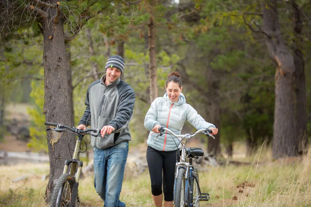 Two individuals walk their bicycles along a tree-lined path surrounded by greenery, suggesting easy access to nearby parks and nature trails. The mood is joyful and adventurous, highlighting exploration and active leisure in the area.