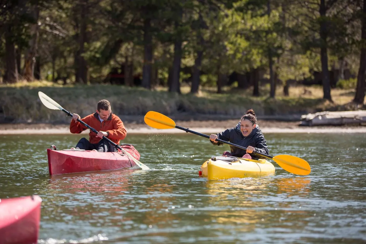 Two people paddle brightly colored kayaks across a shimmering lake, surrounded by nature. The scene is vibrant and full of energy, evoking a sense of adventure and outdoor fun.