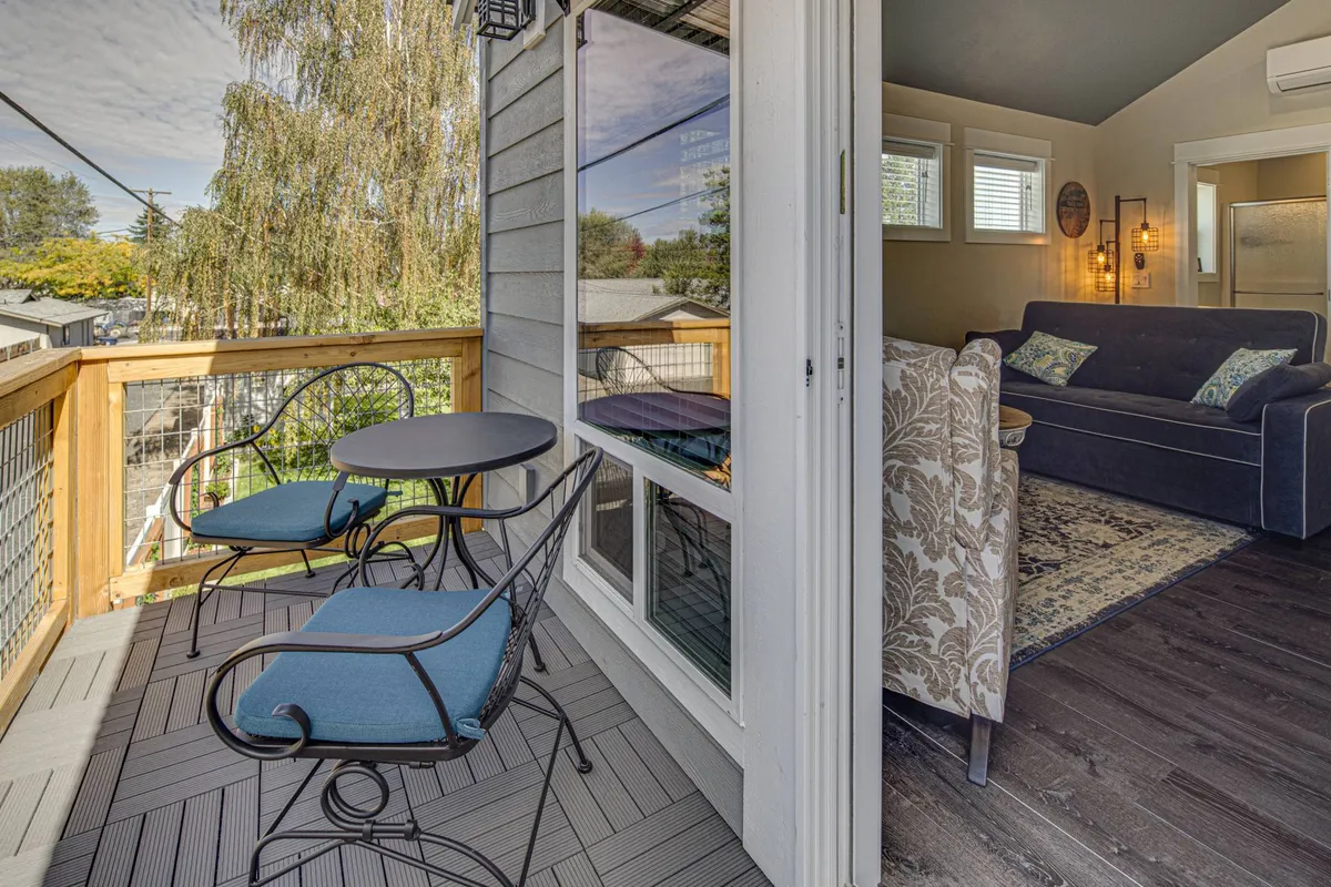 An exterior shot of a cozy deck nook with a small round table and two chairs, overlooking a large window and with a glimpse of the living area inside. This intimate corner invites morning coffee, peaceful reading, or intimate conversation in the fresh air.