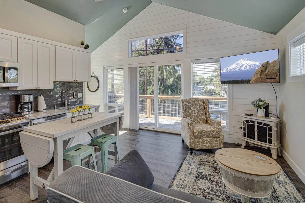 The image showcases a bright, open living area featuring a kitchen counter and a cozy dining table set beside large windows. Sunlight pours in, illuminating comfortable seating and a smart TV displaying a scenic mountain view. The atmosphere is warm, inviting, and ideal for relaxing or enjoying meals together.