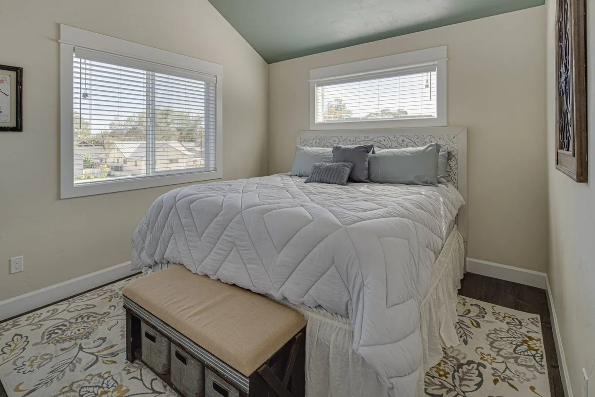 A calm and restful bedroom with a king-size bed featuring a decorative headboard, a bench at the foot of the bed, a decorative rug, and large windows with blinds. The room is sunlit, evoking a sense of comfort and tranquility.